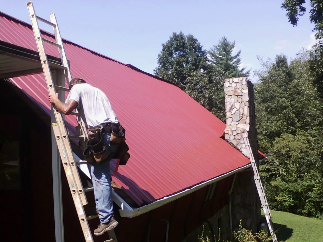 Roofer on a ladder beside a red metal roof and stone chimney — Soffit & Fascia Repair by Roofing Brothers in Spring Arbor